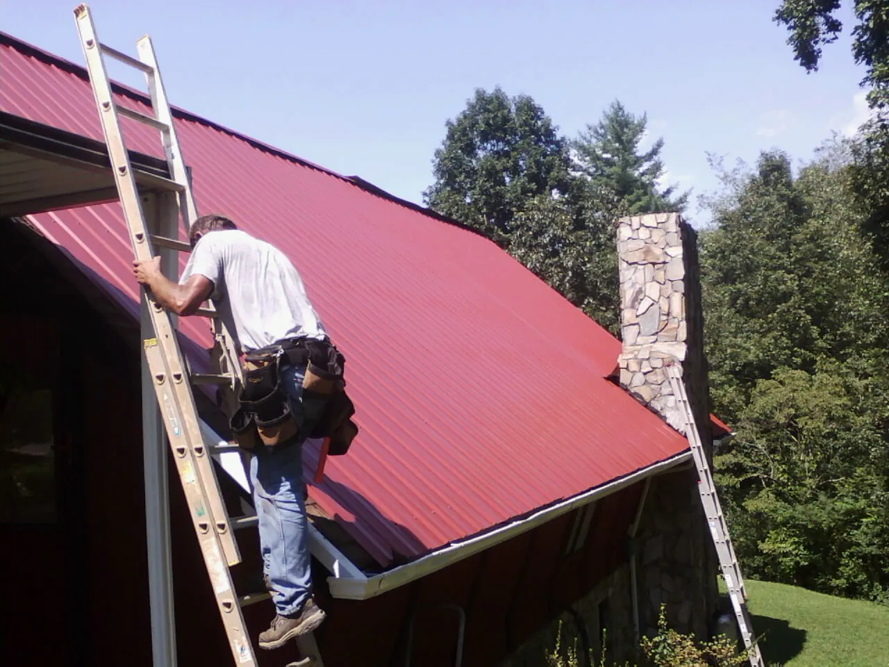 Roofer on a ladder beside a red metal roof and stone chimney — Soffit & Fascia Repair by Roofing Brothers in Larimer